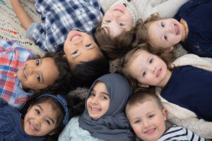 A multi-ethnic group of kids is laying on the carpet of their preschool classroom. They are smiling up at the camera while posing for a picture.